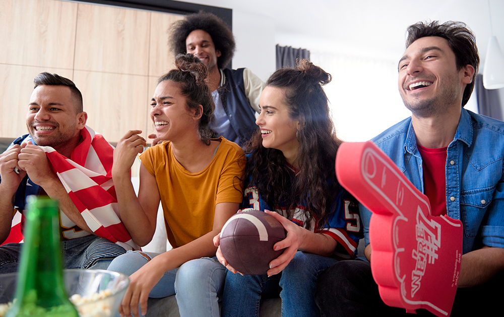 Group of friends watching a football game together on a couch with snacks and fan gear during a gameday gathering in a rental home