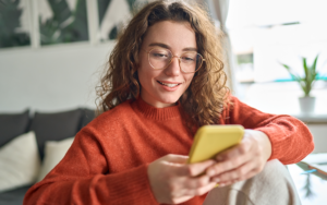 A person sits on a sofa indoors, holding a yellow smartphone, wearing a rust-colored sweater and round glasses in a softly lit living space.