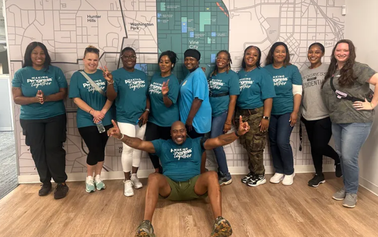 Group of eleven people wearing matching teal “Making Progress Together” T-shirts pose in front of a large wall map graphic in an office setting.