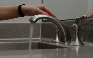 Hand turning on a bathroom sink faucet with running water, representing routine drain use and potential clogs in a rental home
