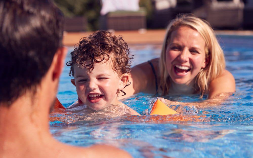 A smiling young child wearing orange arm floaties swims in a pool while two adults closely supervise nearby, demonstrating active water safety and family fun during the summer.