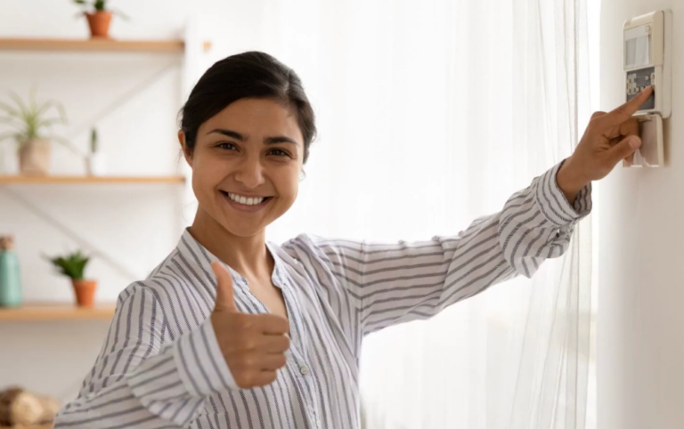 A woman stands beside a wall-mounted thermostat, pressing the controls with one hand and giving a thumbs-up with the other. The room is brightly lit with natural light, and decorative shelves with potted plants are visible in the background.