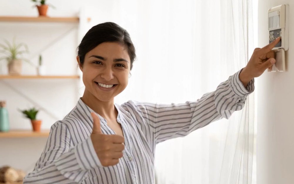 A woman stands beside a wall-mounted thermostat, pressing the controls with one hand and giving a thumbs-up with the other. The room is brightly lit with natural light, and decorative shelves with potted plants are visible in the background.