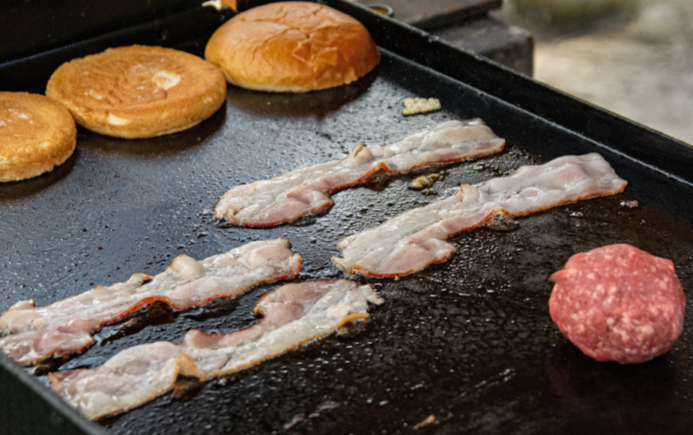 Several strips of bacon cook on a flat-top griddle alongside burger buns and a ball of raw ground beef, with grease glistening on the hot cooking surface.