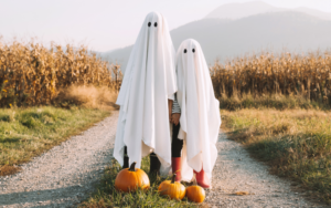 Two people wearing simple white sheet ghost costumes stand on a gravel path beside several pumpkins, with a cornfield and distant mountains in the background on an autumn day.