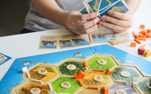 Person arranging pieces and resource cards while playing a strategy board game on a tabletop, with colorful game tiles, dice and wooden game pieces visible.